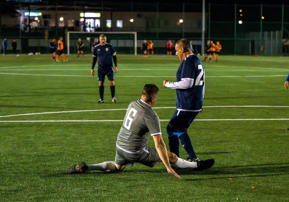 Das Foto zeigt eine nächtliche Szene auf einem Kunstrasen-Fußballfeld, das trotz der dunklen Umgebung hell erleuchtet ist. Der Fokus liegt auf zwei Fußballspielern im Vordergrund, die sich in einem dynamischen Spielmoment befinden. Ein Spieler, ein graues Trikot mit der Nummer 8, schlittert über den Kunstrasen, scheinbar getackelt oder nach einem umkämpften Ball fallend. Seine Haltung zeugt von Anstrengung und vielleicht auch leichter Frustration. Der zweite Spieler, ein dunkelblaues Trikot (Nummer teilweise sichtbar, aber vermutlich im 20er-Bereich), positioniert sich über dem grauen Spieler, als hätte er gerade getackelt oder kämpft um den Ball. Seine Körpersprache zeugt von Dringlichkeit und Konzentration. Die weißen Socken beider Spieler heben sich von den dunklen Trikots und dem grünen Spielfeld ab. Im Hintergrund sind mehrere weitere Spieler zu sehen, teils in Bewegung, teils stillstehend, was auf ein laufendes Spiel schließen lässt. Sie sind unscharf und weniger scharf, was den Blick zurück auf das zentrale Geschehen lenkt. Im Hintergrund sind ein teilweise sichtbarer Torpfosten und ein Schiedsrichter in einem leuchtend orangefarbenen Trikot zu sehen, was das Bild zusätzlich als Fußballspiel kontextualisiert. Die Atmosphäre ist geprägt von intensivem Wettkampf im Kontext eines Amateur- oder Freizeitspiels. Im Hintergrund sind einige Zuschauer zu sehen. Die Gesamtstimmung ist von Action und Konzentration geprägt, mit einer leicht rauen und realistischen Darstellung eines aktiven Fußballspiels. Die Beleuchtung betont die Spieler im Vordergrund und erzeugt ein Gefühl von Dramatik und Unmittelbarkeit. Der Kunstrasen ist deutlich sichtbar und trägt zum modernen und vielleicht etwas weniger formellen Ambiente des Spiels bei.